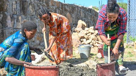 Three women working together to mix concrete in a construction area under bright sunlight.