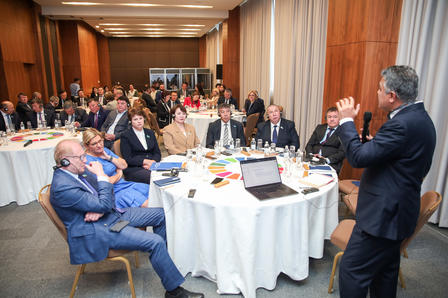 A speaker addresses a seated audience at a conference, with various attendees engaged.