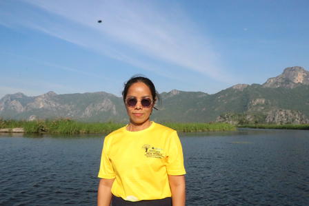 Woman in yellow shirt and sunglasses stands by a lake with mountains in the background.