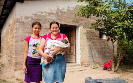 Two women stand outside a clay house, one holding a wrapped baby, surrounded by greenery.