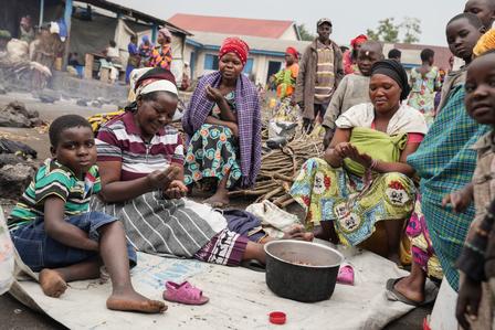 A group of people gathered around a cooking pot outdoors, engaging in conversation and activities.