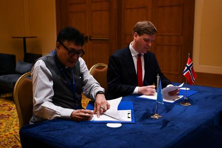 Two men signing documents at a table with flags of Norway and another country between them.