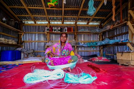 A woman in colorful attire sitting on a red mat, surrounded by yarn and tools in a workshop.