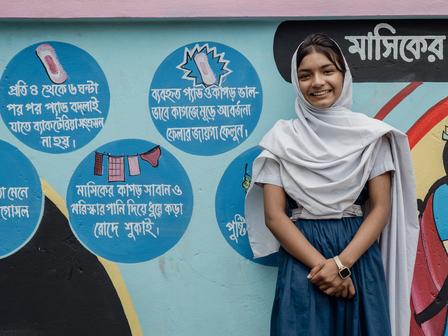 Young woman in a hijab smiles in front of a colorful educational wall mural.