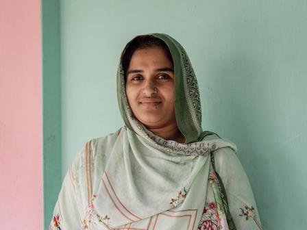 Smiling woman in a light green headscarf, standing against a pastel wall.