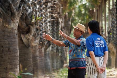 A man in a hat gestures while talking to a woman in a blue shirt among palm trees.