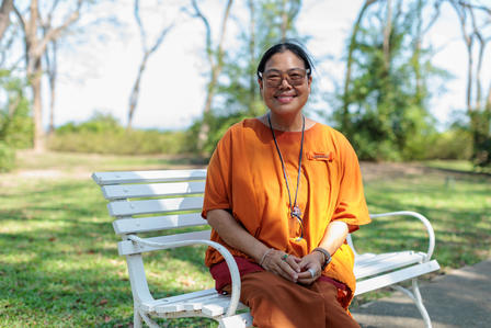 Smiling woman in an orange top sitting on a bench in a park with greenery and trees.