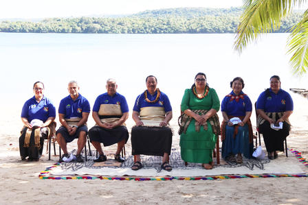 Eight individuals seated on a beach, dressed in traditional attire, with greenery and water in the background.