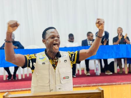 A man with raised arms passionately speaks at a podium, surrounded by seated attendees.