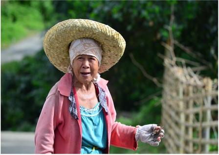 Elderly woman in a straw hat and colorful clothing, smiling in a rural setting.