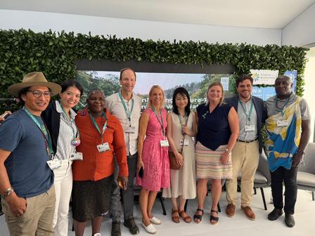 A diverse group of nine people stands together, smiling, in front of a green wall.