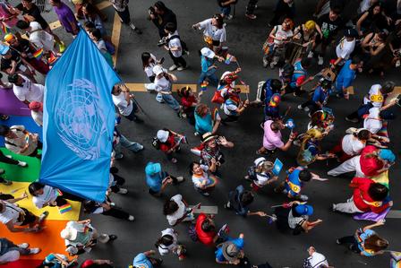 Aerial view of a diverse crowd at a parade, featuring a blue UN flag and colorful road decorations.
