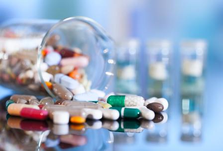 Scattered capsules and tablets next to a tipped-over glass jar, with blurred vials in the background.