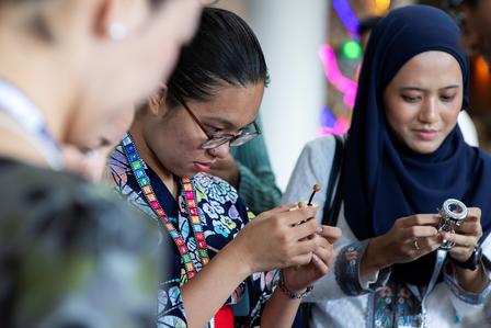 Two women, one wearing a hijab, focus on crafting with tools against a colorful backdrop.