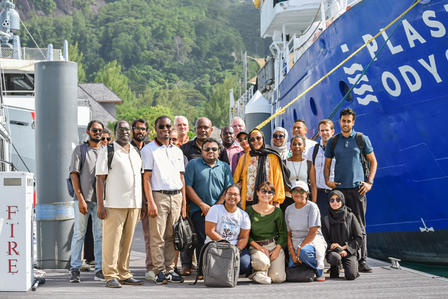 Group of diverse individuals posing on a dock near a blue ship, surrounded by greenery.