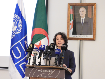 A woman speaks at a podium with microphones, flags, and a portrait in the background.
