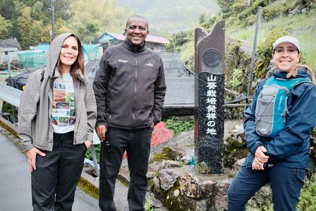 Three people stand near a landmark in a lush outdoor setting, smiling at the camera.