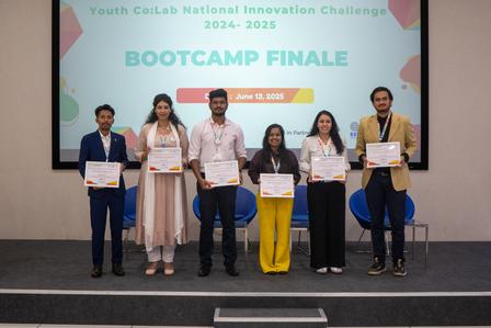 Six participants holding certificates stand on stage in front of a colorful banner.