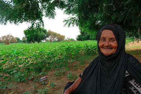 Elderly woman in black attire smiling, seated near green fields and trees.
