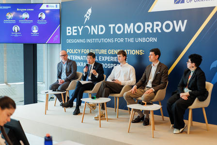 Panel discussion with five speakers seated on stage, backdrop features "Beyond Tomorrow" text.