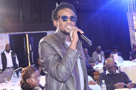 A man in sunglasses performs with a microphone in a well-lit event space.