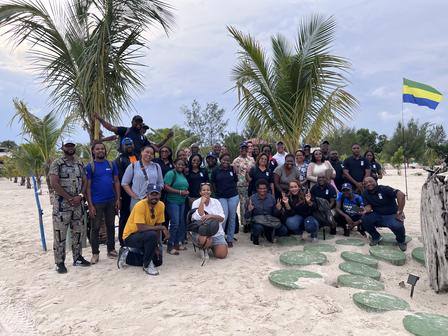 Group of people posing together on a sandy beach with palm trees and a flag in the background.