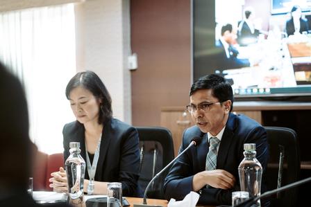Two professionals at a conference table, engaged in discussion, with a video call visible behind them.