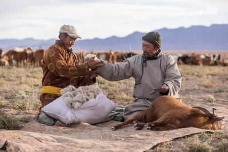 Two men sit on the ground, exchanging items, surrounded by livestock and mountains.