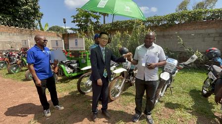 Two men shake hands under a green umbrella, with motorcycles in the background.