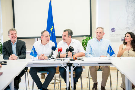 A group of five people at a table during a press conference, with a blue flag in the background.
