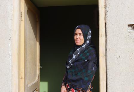 A woman in a patterned headscarf stands in the doorway of a building, smiling.