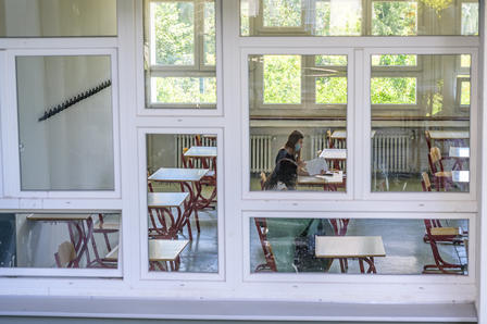 A student sitting at a desk inside a classroom, visible through large windows.