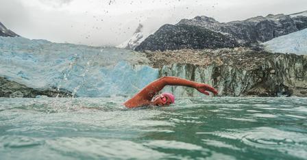 A swimmer in a pink cap strokes through cold water near a glacier, surrounded by mountains.