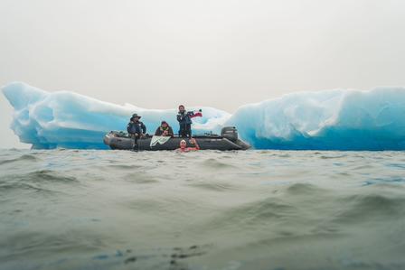 Three people in a small boat near a large blue iceberg on a foggy day.