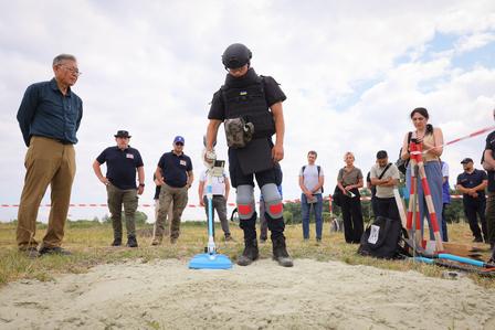 A group of people watches a man using a detection tool on the ground outdoors.