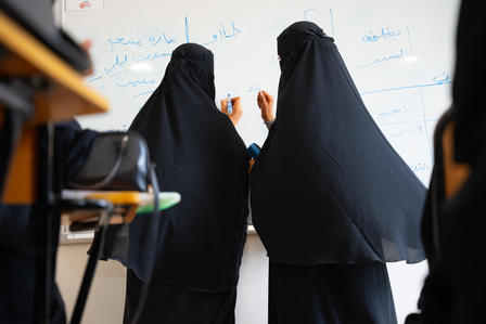 Two women in black abayas write on a whiteboard in a classroom setting.