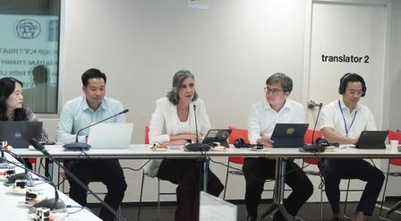A group of professionals seated at a table during a meeting, with laptops and a translator booth.