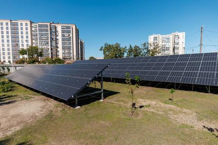 Solar panels arranged in rows on a grassy area, with buildings in the background.