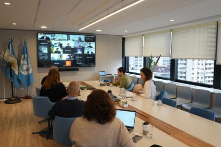 Un grupo de personas en una sala de reuniones moderna, participando de una videollamada proyectada en una pantalla.