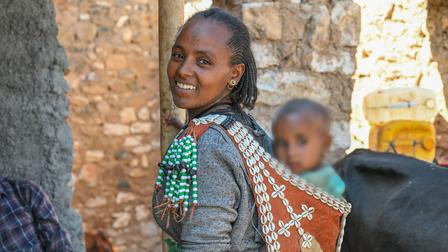 Smiling woman in traditional attire with child on her back, in a rustic setting.