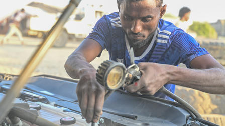 A man in a blue shirt works on a car engine with a tool, focused and engaged in repairs.