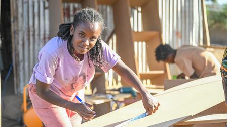 A girl measures wood with a pencil, smiling, while another person works in the background.