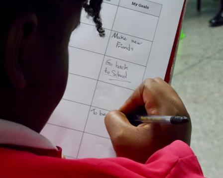 A child's hand holding a pen, writing on a sheet of paper with a red folder.
