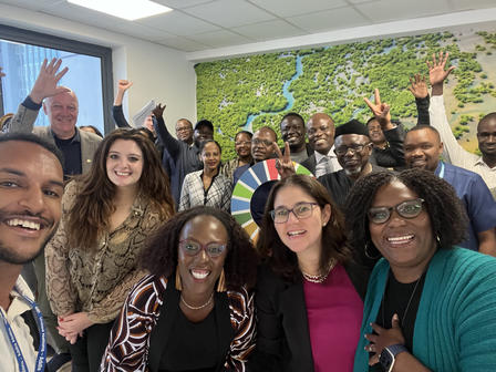 A diverse group of people smiling and posing in a bright office with a green mural backdrop.