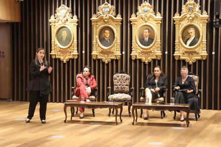 A speaker addresses an audience while three panelists sit on stage with ornate portraits behind them.