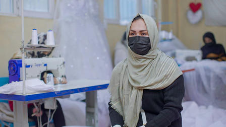A woman in a headscarf sits in front of a sewing machine, focused and wearing a mask.