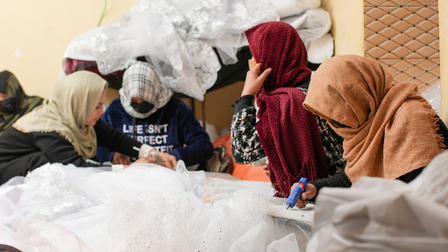 Four women, wearing headscarves, closely work together on fabric in a bright, cluttered room.