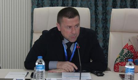 A man in a dark suit speaks at a conference table, with water bottles and documents visible.