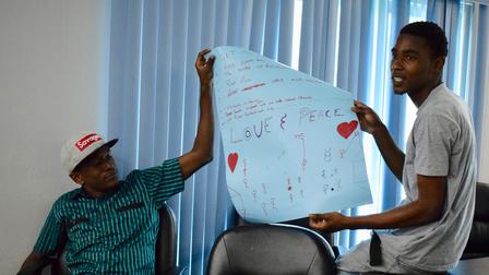 Two young men in a room holding a blue poster with red hearts, promoting love and peace.