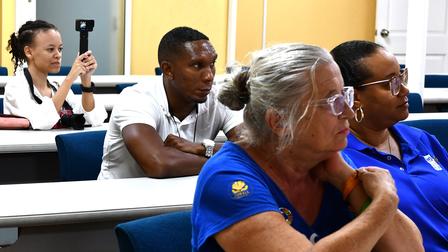 A group of people seated in a classroom, attentively listening, with varied expressions.